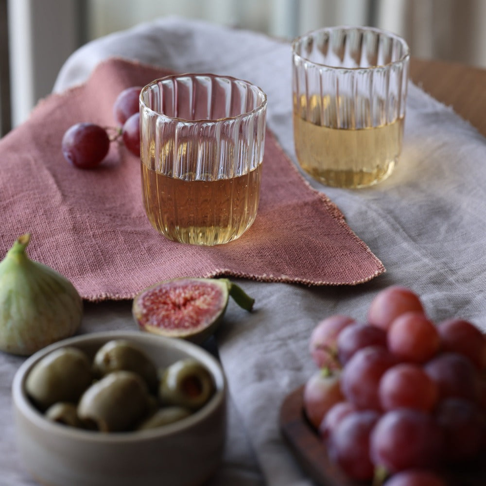 Two glasses of amber liquid on a pink cloth with figs, olives, and grapes on a table.