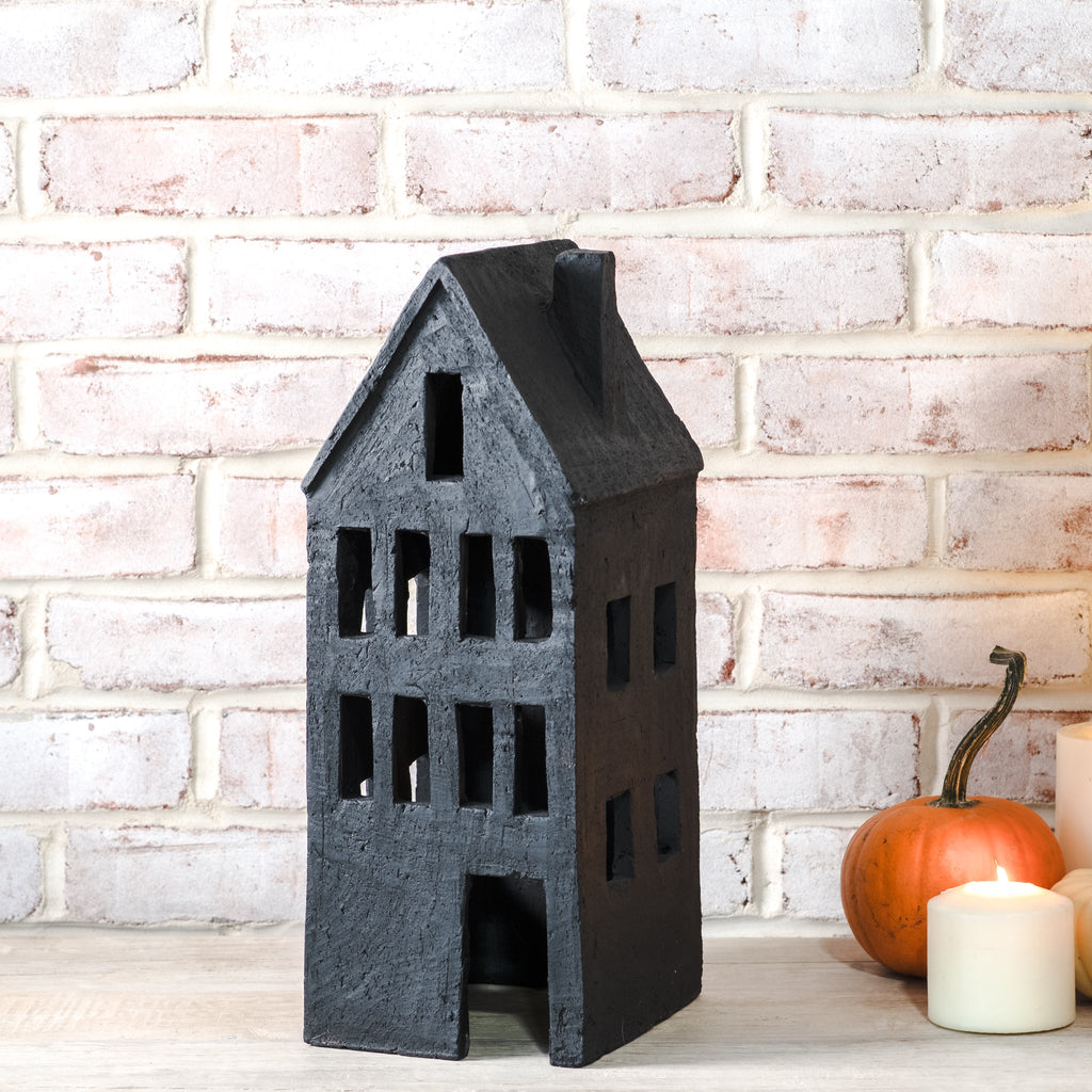A black decorative house-shaped candle holder with cutout windows, displayed on a light wooden surface against a white brick wall, with a small orange pumpkin and white candles as props.
