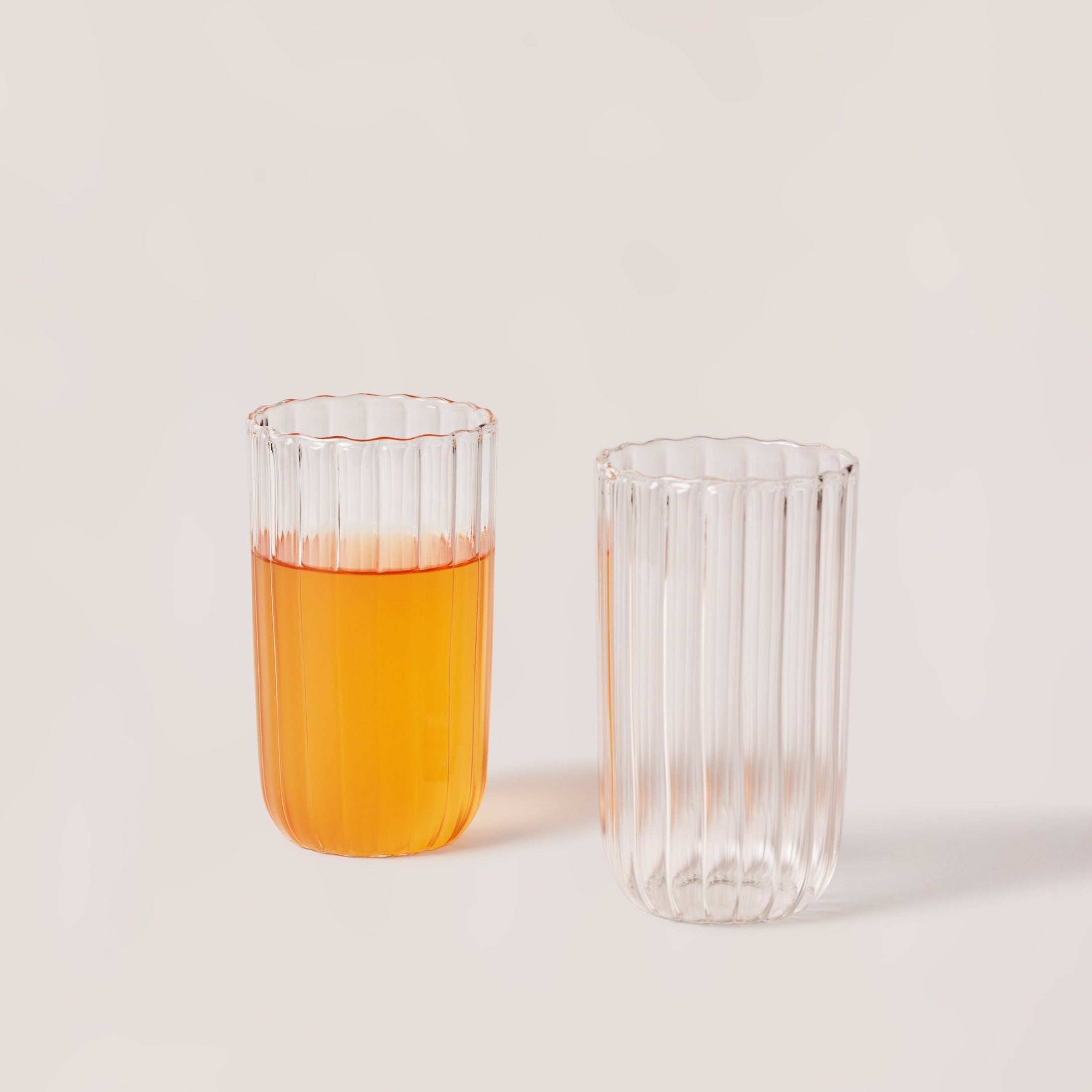 Two glass tumblers, one filled with orange juice, on a light background