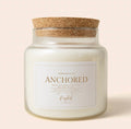 A glass jar candle with white wax and a natural cork lid, featuring a white label with gold lettering that reads 'ANCHORED' and 'Raspberry Creek Candle Co.' on a light beige background.