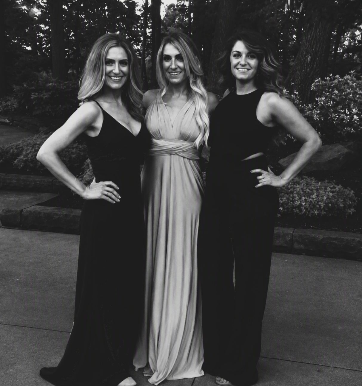 Three women posing together outdoors in elegant formalwear. The woman on the left wears a sleeveless black gown, the center woman wears a light-colored wrap-style maxi dress, and the woman on the right wears a black sleeveless jumpsuit.