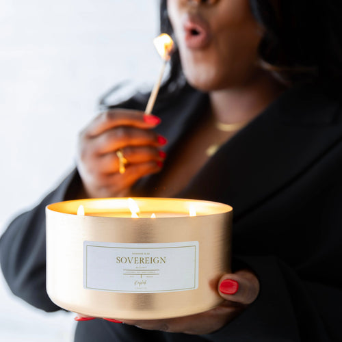 A person in a black blazer holds a lit gold candle jar labeled 'SOVEREIGN' while lighting a match with a matchstick. The person has red nail polish and is wearing a gold ring, set against a bright, softly blurred background.