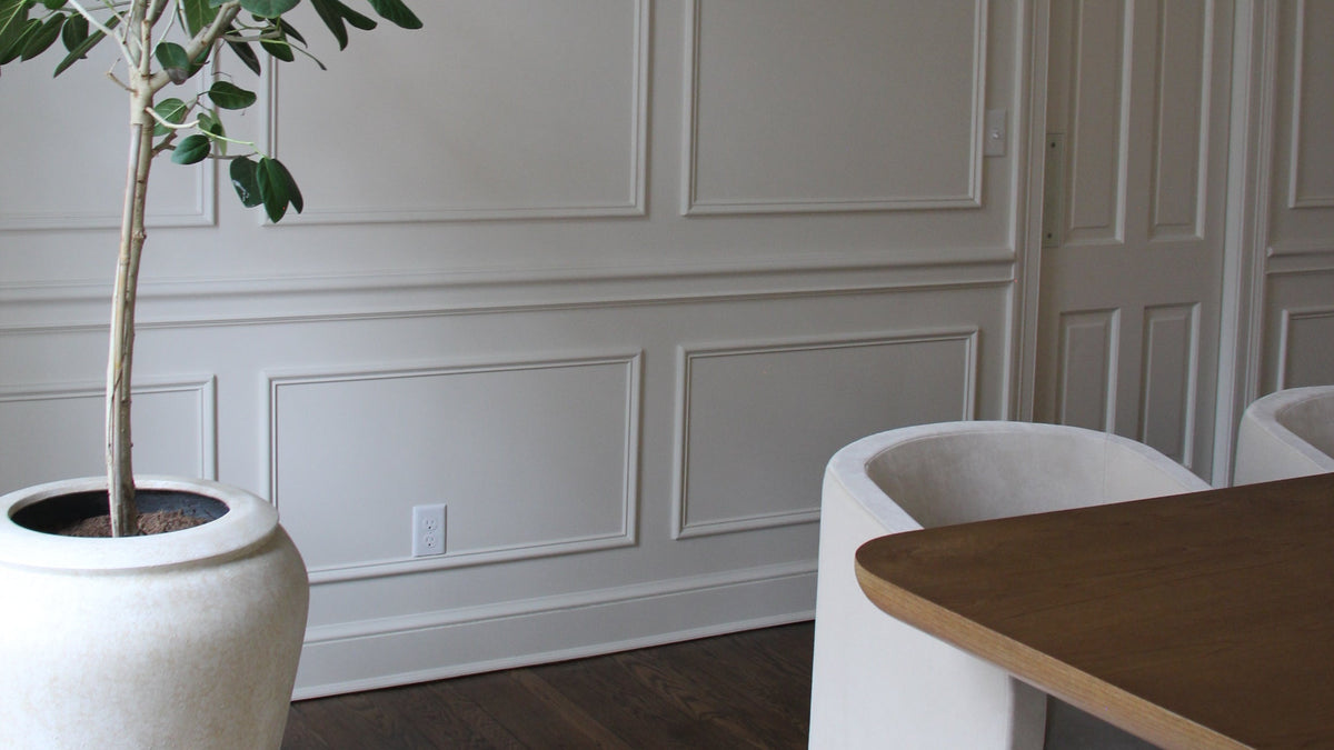 A large potted tree with green leaves in a textured off-white ceramic pot, placed in a modern room with white paneled walls, a wooden dining table, and white chairs.