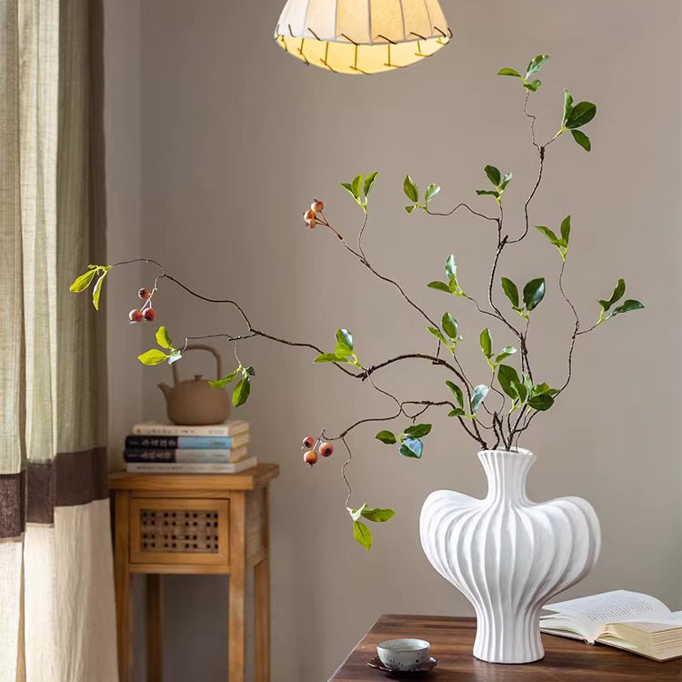 White vase with decorative branches on a wooden table against a beige wall.