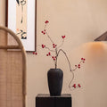 Black vase with red branches on a table against a beige wall with a painting.