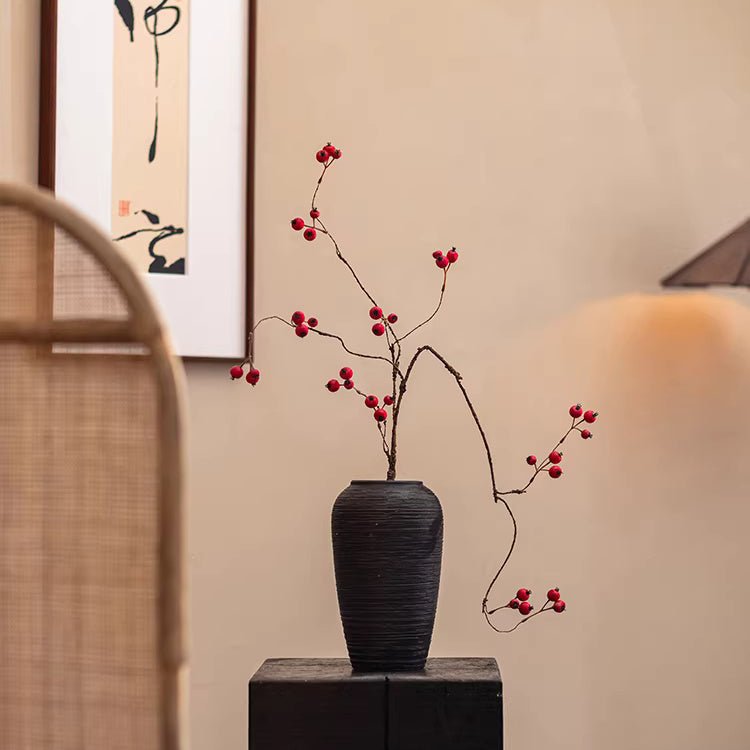 Black vase with red branches on a table against a beige wall with a painting.