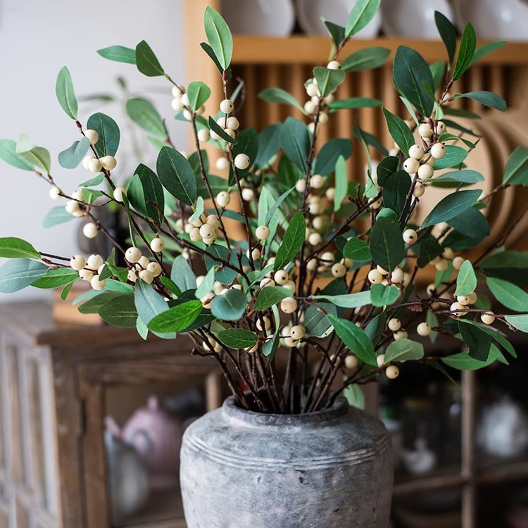 Faux greenery with small white berries in a gray vase on a blurred indoor background