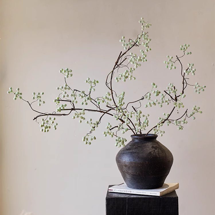 Decorative arrangement of branches with small green berries in a dark vase on a wooden surface.