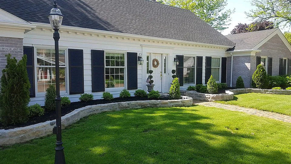 Front exterior of a single-story house with white siding, dark shutters, a dark roof, a green lawn, landscaped garden beds, and a black decorative lamp post in the foreground.