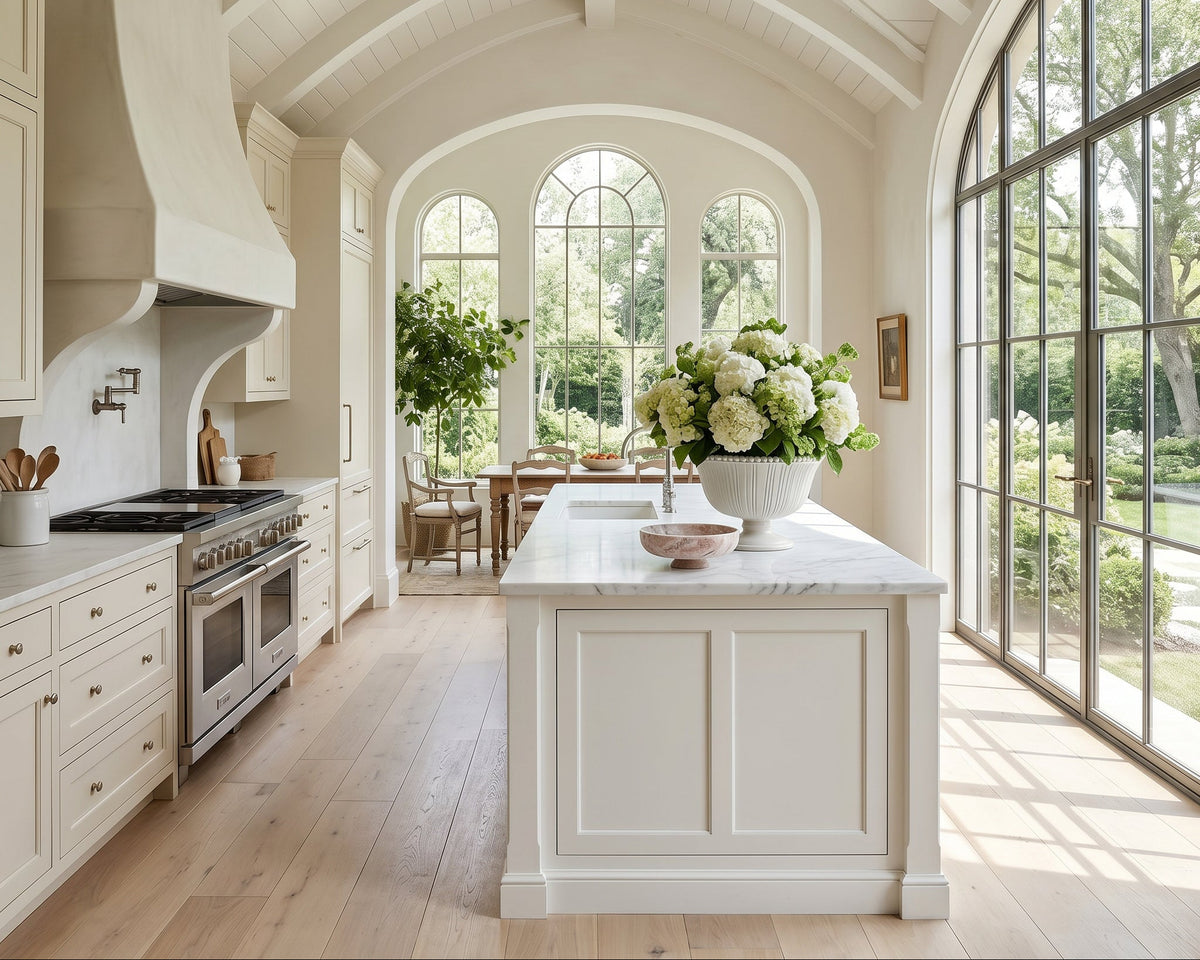 A bright, elegant kitchen interior with white cabinetry, a marble island, a large floral centerpiece, and tall arched windows looking out to a garden. No people or animals are present.