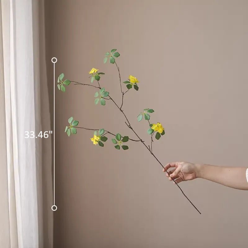Hand holding a decorative branch with yellow flowers against a beige wall.