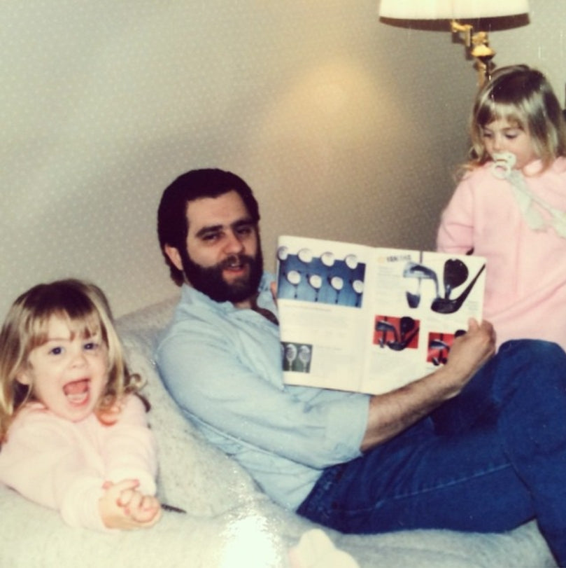 A vintage-style photo shows a man with a beard sitting on a couch with two young girls. The man is wearing a light blue button-up shirt and jeans, holding an open magazine or book.