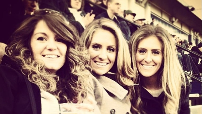 Three women smiling and posing together in crowded stadium seating, with a warm sepia-toned filter applied to the photo. One woman holds a drink cup.