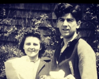 A vintage black and white photograph showing a smiling woman holding a paper bag and a man in a vest holding a white bag, standing outdoors in front of a stone wall.