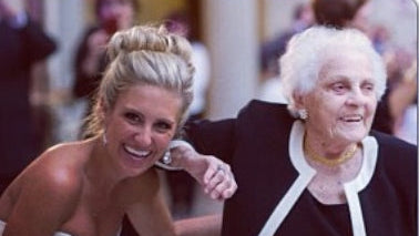 A smiling blonde woman in a white strapless dress leans in beside an older woman with white hair wearing a black outfit with white trim, in a crowded indoor setting.