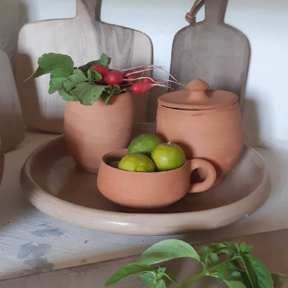 Terracotta kitchenware including a pot, bowl, and cup on a wooden surface with green apples and radishes.