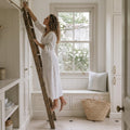 Woman in a white dress standing on a wooden ladder in a bright kitchen with large windows.