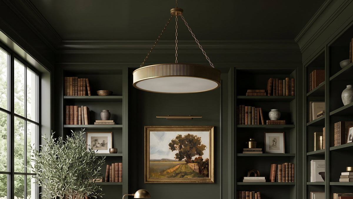A styled home office interior featuring dark green built-in bookshelves, a dark wood writing desk, a cream upholstered armchair, a large patterned area rug, a hanging pendant light, and a framed landscape painting on the wall.