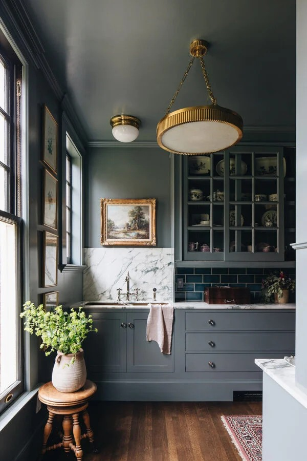 A styled interior kitchen scene with dark blue-gray cabinets, a white marble backsplash, dark subway tile, a hanging brass pendant light, wood floors, and a vase of greenery on a small wooden stool.