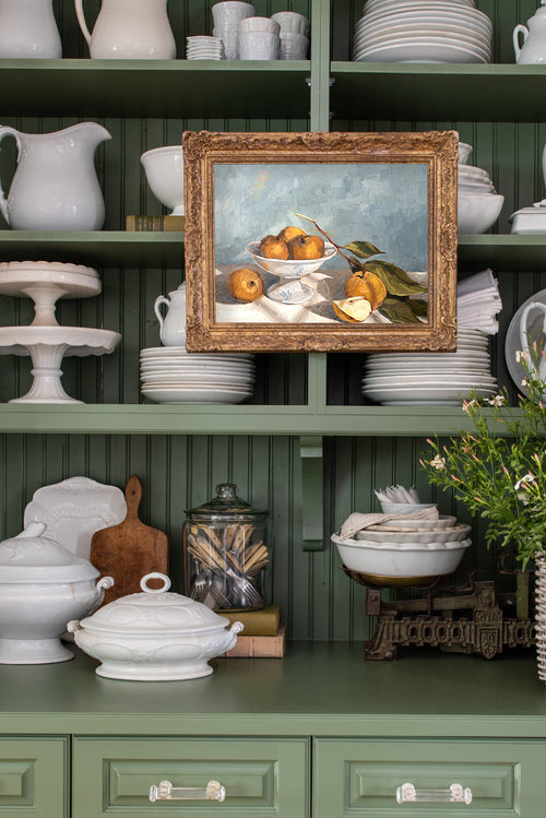 Green kitchen shelf with white ceramic items and a framed painting of fruit.