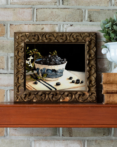Framed still life painting of grapes on a table against a brick wall.