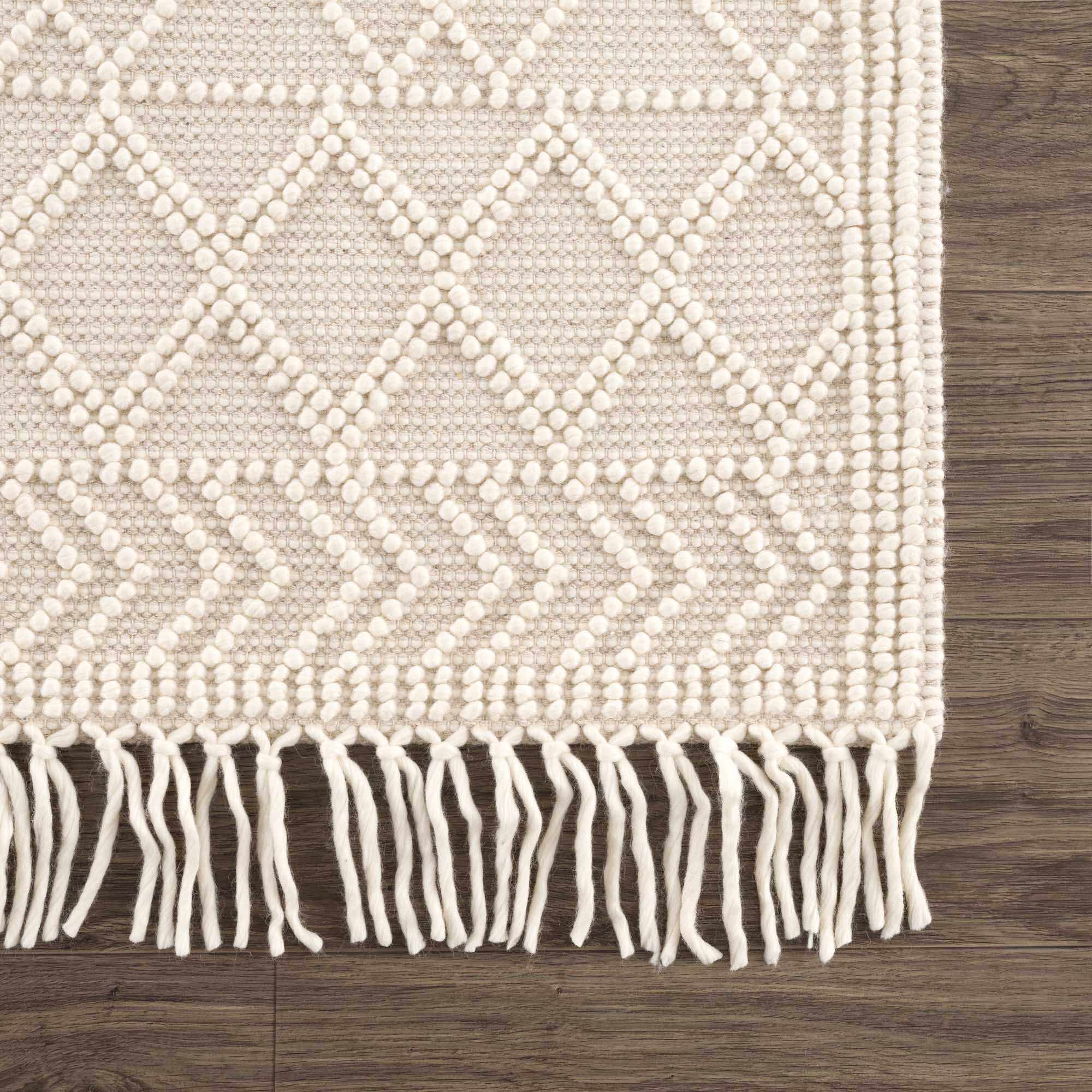 Beige woven rug with diamond pattern and white fringe on a wooden floor.