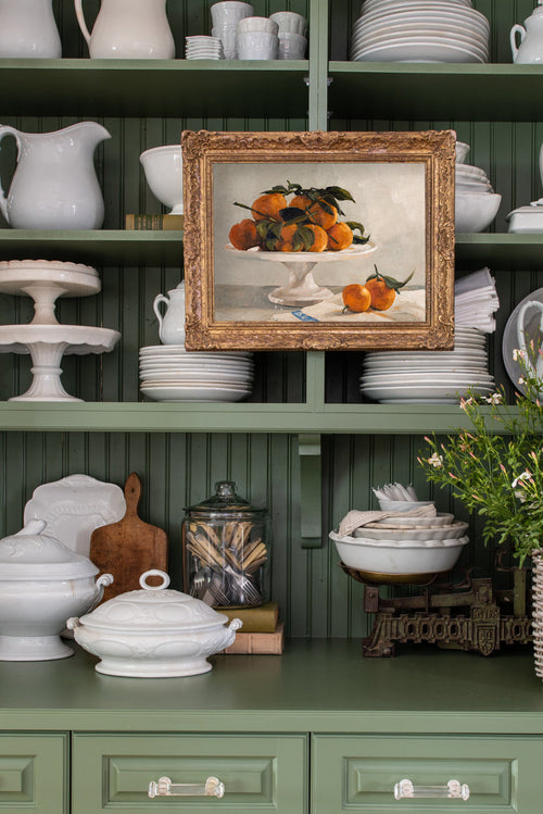 Green kitchen shelf with white ceramic dishes, a framed painting of oranges, and decorative items.