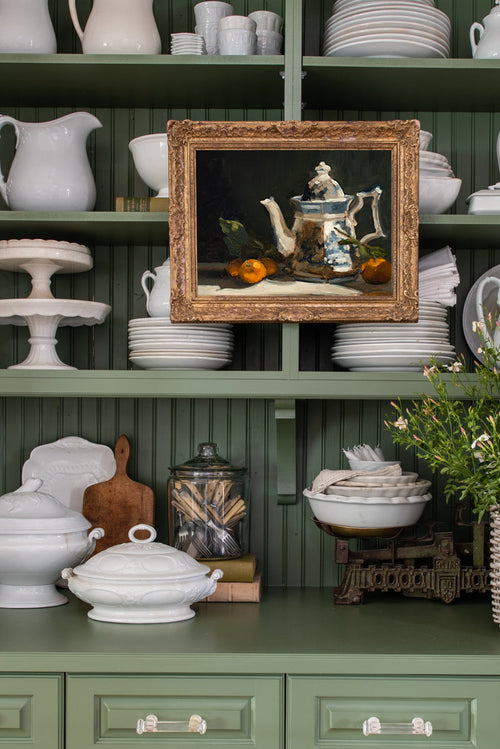 Green kitchen cabinet with white dishes, a framed picture, and decorative items.
