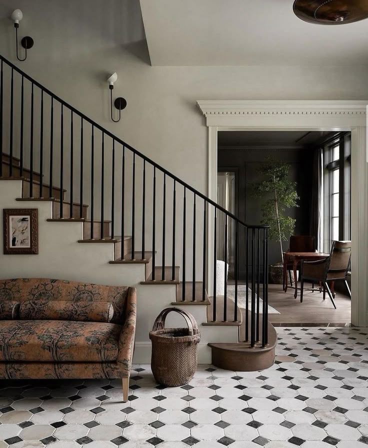 A styled interior entryway featuring a staircase with black metal railings and wooden steps, a patterned floral sofa, a basket on a black-and-white diamond-tiled floor, and an open doorway leading to a dining area.