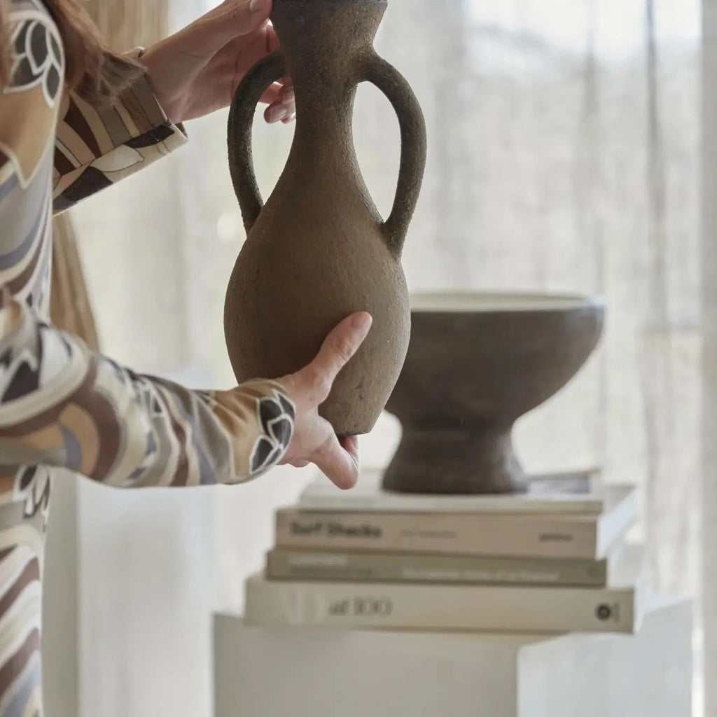 Person holding a brown ceramic vase with another vase in the background on a surface with books.