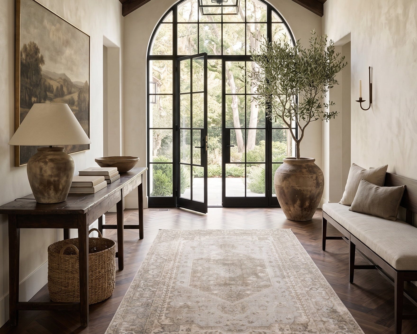 A long, elegant entryway or hallway with a high wood-beamed vaulted ceiling, dark wood floors, a large beige patterned area rug, a console table with a lamp, a bench with cushions, and a large potted tree near arched glass doors.
