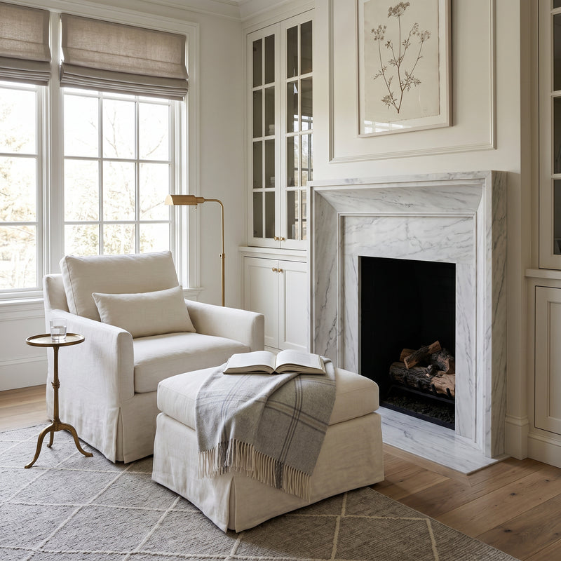 A styled living room corner featuring a cream slipcovered armchair with a matching pillow, a cream ottoman draped with a gray plaid throw and an open book, a small gold side table, a floor lamp, a white marble fireplace, and a patterned rug.